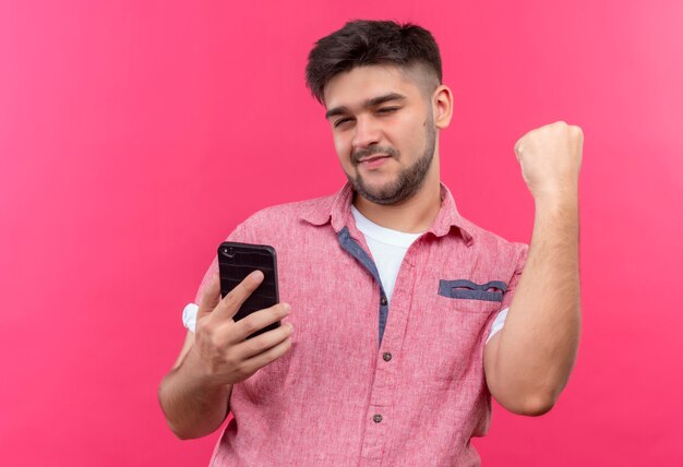 Chico guapo joven con camisa polo rosa parpadeando un ojo sosteniendo el teléfono parado sobre la pared rosa