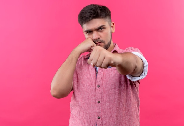 Foto gratuita chico guapo joven con camisa de polo rosa que amenaza airadamente con golpear de pie sobre la pared rosa