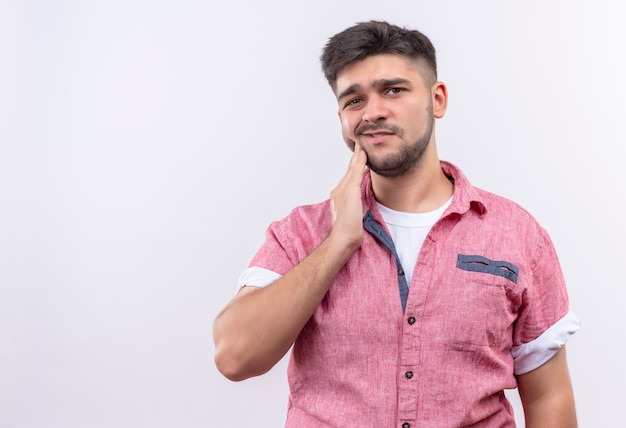 Foto gratuita chico guapo joven con camisa de polo rosa que sufre de dolor de muelas sosteniendo la mejilla con la mano de pie sobre la pared blanca