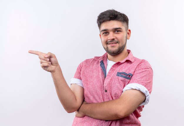 Chico guapo joven con camisa polo rosa sonriendo coridalmente apuntando hacia la derecha con el dedo índice de pie sobre la pared blanca