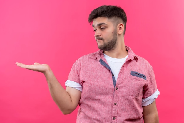 Chico guapo joven con camisa polo rosa sosteniendo algo en su mano izquierda de pie sobre la pared rosa