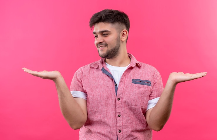 Foto gratuito chico guapo joven con camisa polo rosa sosteniendo algo en sus manos izquierda y derecha mirando la mano derecha de pie sobre la pared rosa