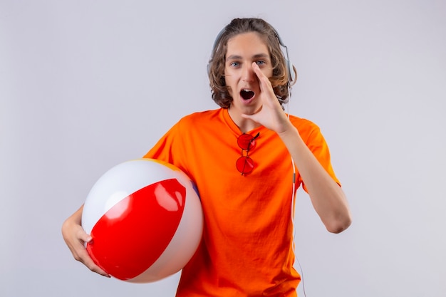 Chico guapo joven en camiseta naranja con auriculares sosteniendo una bola inflable gritando o llamando a alguien con la mano cerca de la boca mirando sorprendido de pie sobre fondo blanco