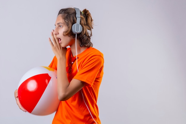 Chico guapo joven en camiseta naranja con auriculares sosteniendo una pelota inflable mirando a un lado diciendo un secreto con la mano cerca de la boca de pie sobre fondo blanco