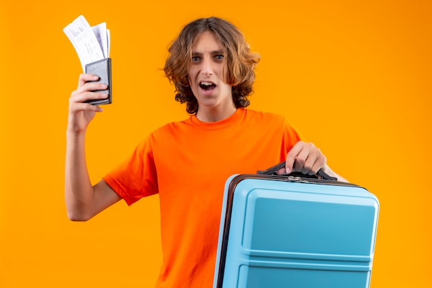 Chico guapo joven en camiseta naranja con billetes de avión y maleta de viaje mirando sorprendido de pie sobre fondo amarillo