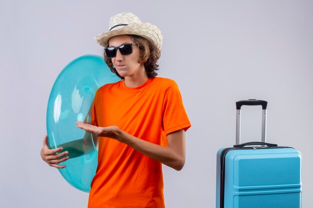 Chico guapo joven en camiseta naranja con gafas de sol negras con anillo inflable mirando confundido presentando con el brazo de la mano de pie con maleta de viaje sobre fondo blanco