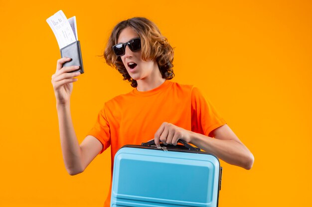 Chico guapo joven en camiseta naranja con gafas de sol negras con billetes de avión y maleta de viaje mirando sorprendido y feliz de pie sobre fondo amarillo