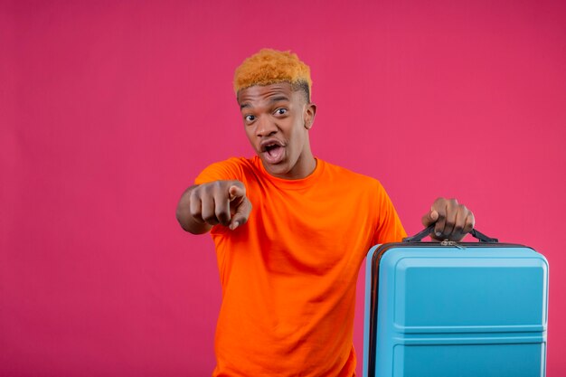 Chico guapo joven con camiseta naranja con maleta de viaje apuntando con el dedo a la cámara mirando sorprendido y asombrado de pie sobre la pared rosa