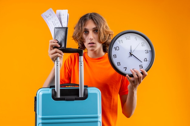 Chico guapo joven en camiseta naranja con maleta de viaje y boletos aéreos de pie con reloj mirando confundido