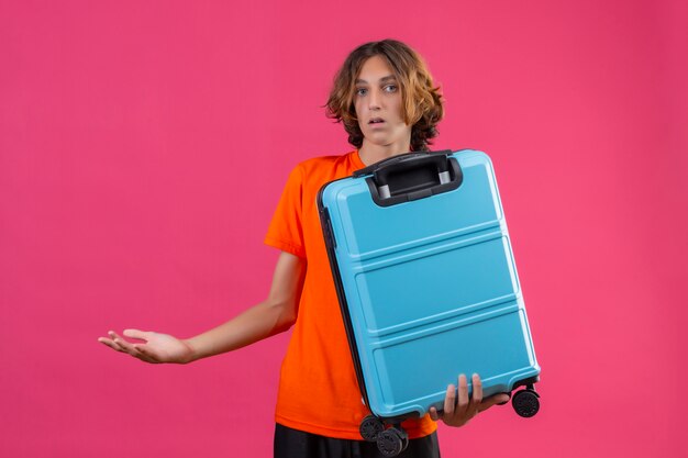 Chico guapo joven en camiseta naranja con maleta de viaje despistado y confundido sin respuesta, extendiendo las manos de pie