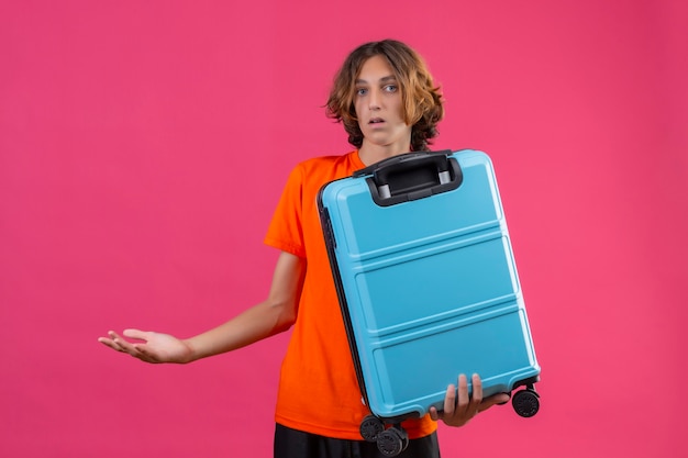 Foto gratuita chico guapo joven en camiseta naranja con maleta de viaje despistado y confundido sin respuesta extendiendo las manos sobre fondo rosa