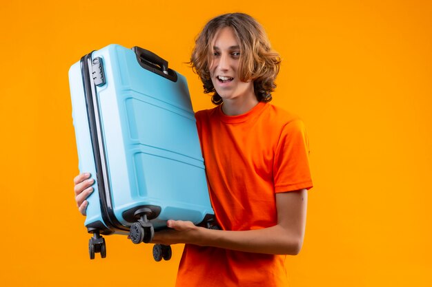 Chico guapo joven en camiseta naranja con maleta de viaje positiva y feliz sonriendo alegremente de pie sobre fondo amarillo