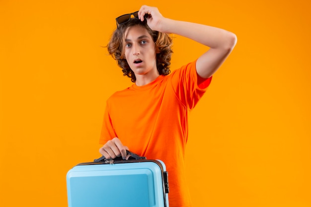 Chico guapo joven en camiseta naranja con maleta de viaje quitándose las gafas mirando sorprendido y asombrado de pie