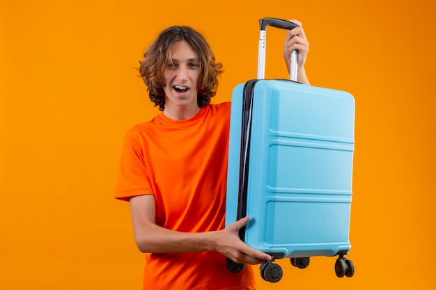 Chico guapo joven en camiseta naranja con maleta de viaje sonriendo alegremente positivo y feliz listo para viajar de pie sobre fondo amarillo