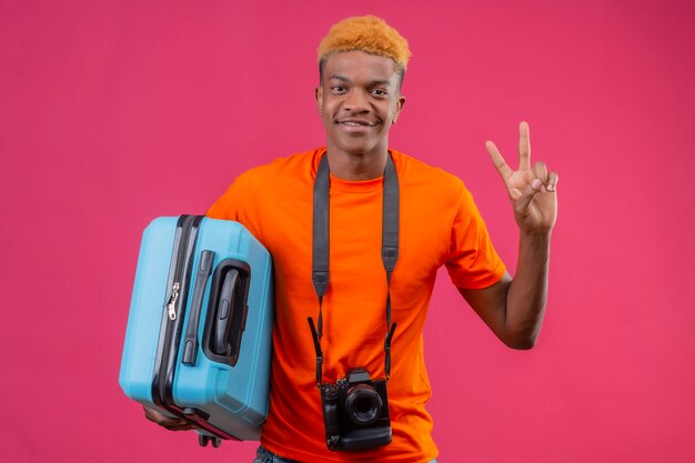 Chico guapo joven con camiseta naranja con maleta de viaje sonriendo amigable mostrando el signo de la victoria