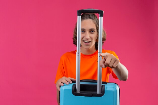 Chico guapo joven en camiseta naranja de pie con maleta de viaje apuntando con el dedo a la cámara sonriendo con cara feliz sobre fondo rosa