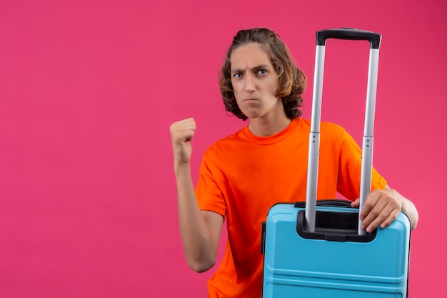 Chico guapo joven en camiseta naranja de pie con maleta de viaje levantando el puño con expresión enojada