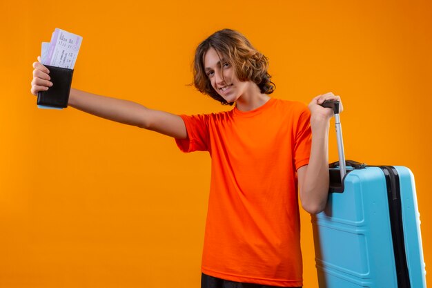 Chico guapo joven en camiseta naranja de pie con maleta de viaje sosteniendo billetes de avión sonriendo alegremente positivo y feliz sobre fondo amarillo