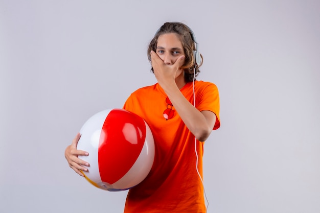 Chico guapo joven en camiseta naranja sosteniendo bola inflable con auriculares asombrado y sorprendido cubriendo la boca con la mano de pie