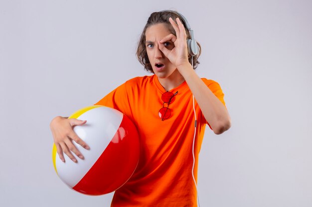 Chico guapo joven en camiseta naranja sosteniendo bola inflable con auriculares haciendo signo ok mirando a través de este signo sorprendido de pie