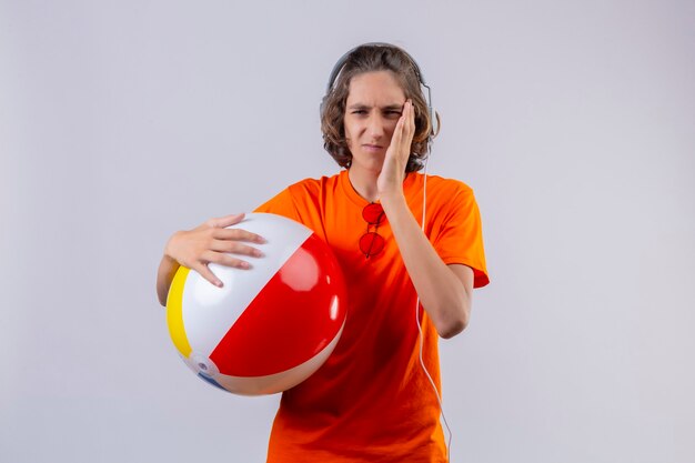 Chico guapo joven en camiseta naranja sosteniendo una pelota inflable con auriculares mirando mal tocar la mejilla con dolor de muelas de pie sobre fondo blanco