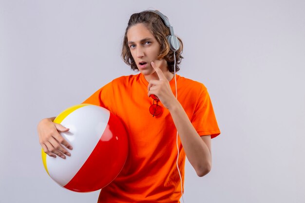 Chico guapo joven en camiseta naranja sosteniendo una pelota inflable con auriculares de pie con mirada pensativa mirando incierto sobre fondo blanco.