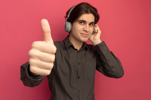 Chico guapo joven complacido con camiseta negra con auriculares mostrando el pulgar hacia arriba aislado en la pared rosa