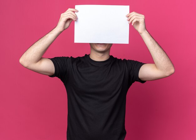 Chico guapo joven complacido con camiseta negra con la cara cubierta con papel aislado en la pared rosa