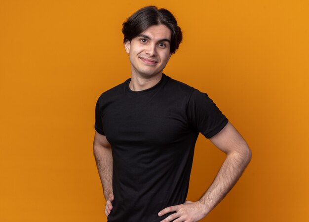 Chico guapo joven complacido con camiseta negra poniendo las manos en la cadera aislada en la pared naranja