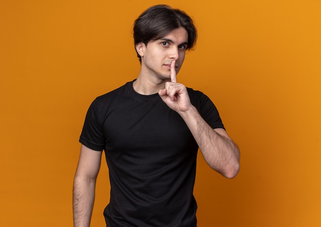 Chico guapo joven complacido con camiseta negra que muestra el gesto de silencio aislado en la pared naranja con espacio de copia