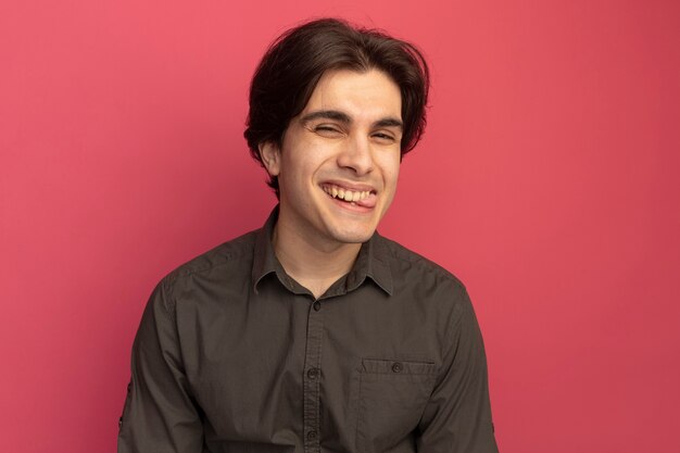 Chico guapo joven complacido con camiseta negra que muestra la lengua aislada en la pared rosa