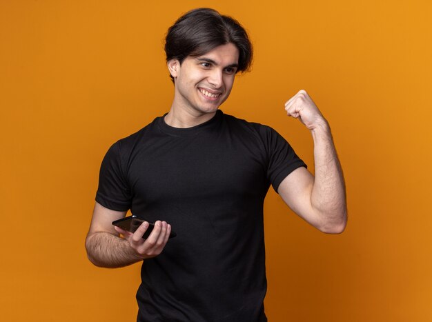 Foto gratuita chico guapo joven complacido con camiseta negra sosteniendo el teléfono y mostrando un gesto de sí aislado en la pared naranja