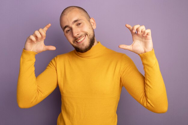 Chico guapo joven complacido fingiendo sostener algo aislado en la pared púrpura