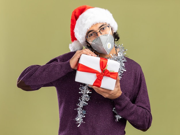 Chico guapo joven complacido con sombrero de navidad y máscara médica con guirnalda en el cuello con caja de regalo aislada sobre fondo verde oliva