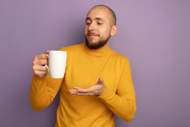 Chico guapo joven complacido sosteniendo y puntos con la mano en una taza de té aislado en la pared púrpura con espacio de copia