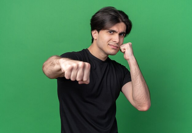 Chico guapo joven confiado con camiseta negra de pie en pose de lucha aislado en la pared verde