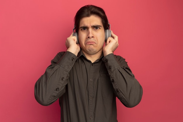 Chico guapo joven confundido con camiseta negra con auriculares aislados en la pared rosa