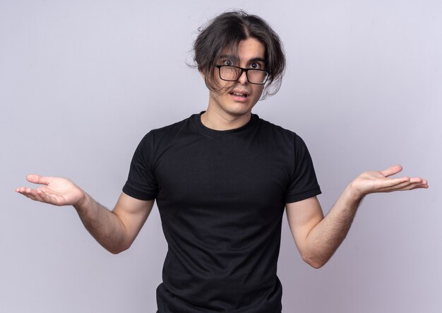 Chico guapo joven confundido con camiseta negra y gafas extendiendo las manos aisladas en la pared blanca