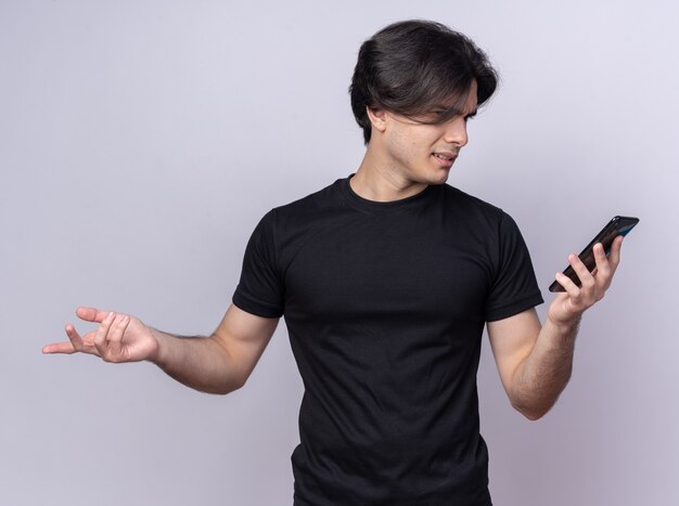 Chico guapo joven confundido con camiseta negra sosteniendo y mirando el teléfono extendiendo la mano aislada en la pared blanca