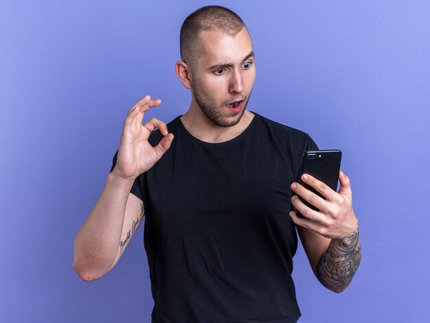 Chico guapo joven confundido con camiseta negra sosteniendo y mirando el teléfono mostrando gesto bien aislado en la pared azul