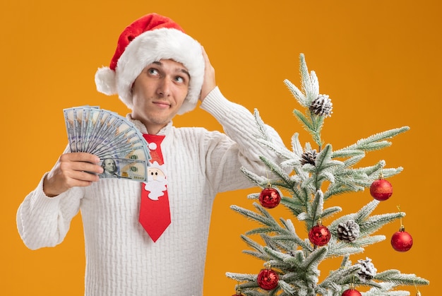 Chico guapo joven confundido con sombrero de navidad y corbata de santa claus de pie cerca del árbol de navidad decorado sosteniendo dinero manteniendo la mano en la cabeza mirando al lado aislado en la pared naranja