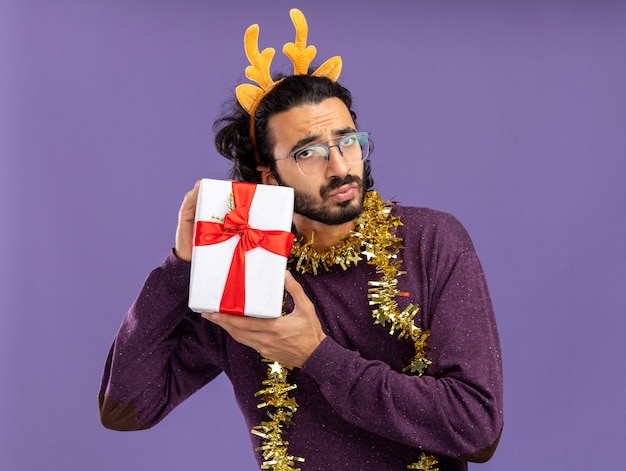 Foto gratuita chico guapo joven disgustado con aro de pelo de navidad con guirnalda en el cuello con caja de regalo aislada sobre fondo azul