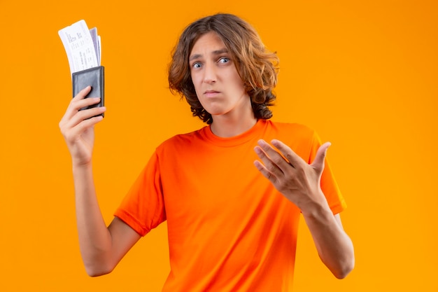 Chico guapo joven disgustado en camiseta naranja con billetes de avión mirando confundido con cara infeliz de pie
