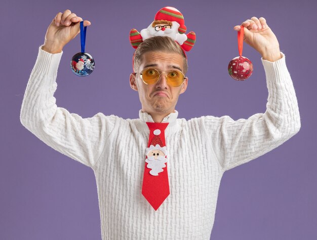 Foto gratuita chico guapo joven disgustado con diadema de santa claus y corbata con gafas levantando adornos de bolas de navidad mirando a cámara aislada sobre fondo púrpura