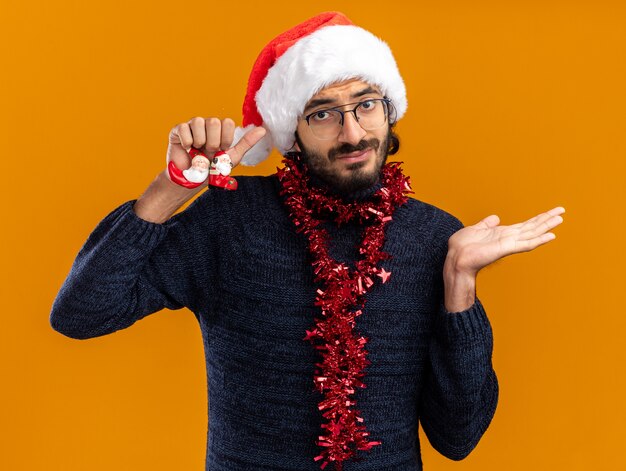 Chico guapo joven disgustado con sombrero de navidad con guirnalda en el cuello sosteniendo juguetes extendiendo la mano aislada sobre fondo naranja