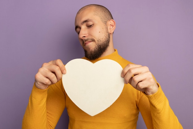 Foto gratuita chico guapo joven disgustado sosteniendo y mirando la caja en forma de corazón aislada en la pared púrpura