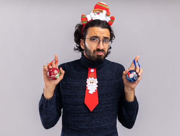 Chico guapo joven disgustado vistiendo corbata de Navidad con aro de pelo sosteniendo bolas de Navidad aislado sobre fondo blanco.