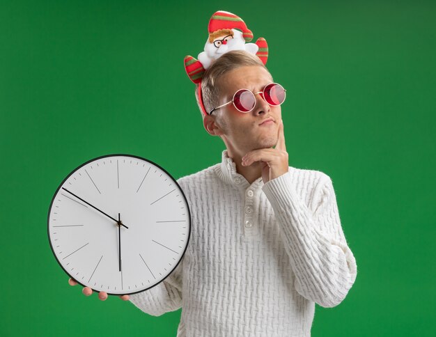 Chico guapo joven dudoso con diadema de santa claus con gafas sosteniendo el reloj manteniendo la mano en la barbilla mirando al lado aislado en la pared verde