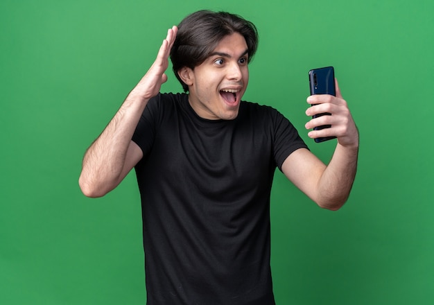 Chico guapo joven emocionado con camiseta negra sosteniendo y mirando el teléfono aislado en la pared verde