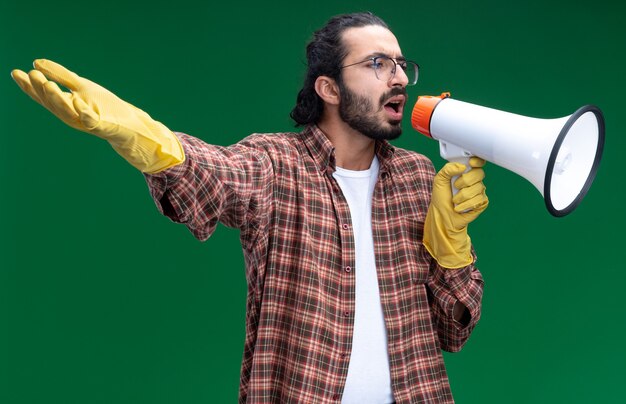 Chico guapo joven emocionado de limpieza con camiseta y guantes habla por altavoz extendiendo la mano aislada en la pared verde